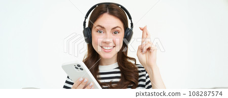 Young woman holding fingers crossed, making wish, hoping for something, holding smartphone, wearing wireless headphones, white background 108287574