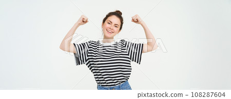 Woman power and feminism. Young girl feeling empowered and strong, flexing her biceps, showing muscles on arms, standing over white background 108287604