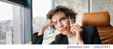 Portrait of woman with frowning face, sitting in office and listening to client over the phone 108287641