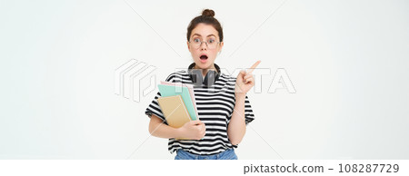 Portrait of girl gasps in surprised, points left with amazed face. Shocked student shows something, pointing finger at banner, holding notebooks, white background 108287729