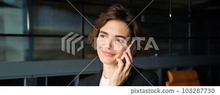 Close up portrait of businesswoman talking on mobile phone, has her smartphone near ear, answers a call, stands in conference room in office building 108287730