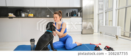Image of young woman does workout from home, sits on yoga mat in living room and plays with her black dog, puppy distracts girl from doing exercises Image of young woman does workout from home, sits on yoga mat in living room and plays with her black dog, puppy distracts girl from doing exercises 108287735