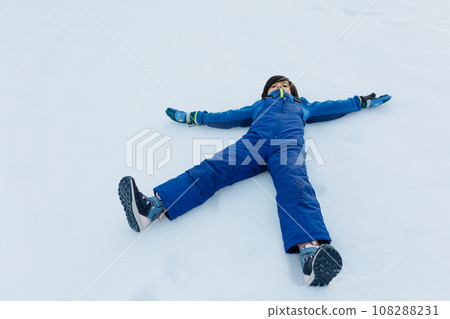 child with dark hair in blue winter pants lying on the snow 108288231