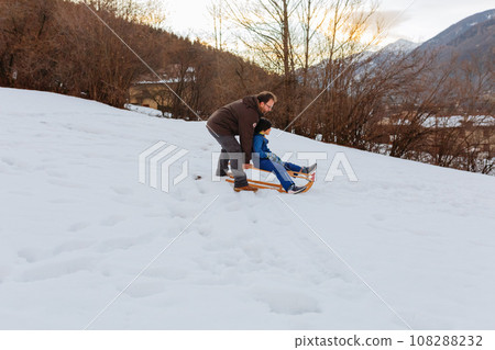 father pushing daughter sitting on wooden sled to slide down the snowy hill 108288232