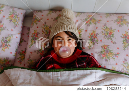 Girl wearing beige warm hat making bubble lying in the bed under blanket 108288244