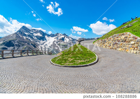 Mountain cobbled road serpentine. Sharp curve with mountain tops on background. High Tauern, Austria Mountain cobbled road serpentine. Sharp curve with mountain tops on background. High Tauern, Austria 108288886