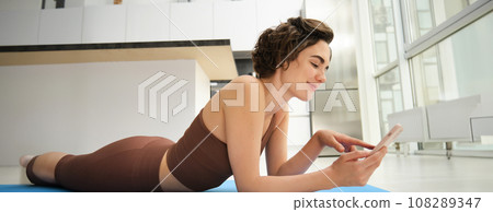 Indoor shot of fit and healthy woman workout at home, lying on yoga mat while using smartphone, watching fitness training at mobile phone, resting after exercises Indoor shot of fit and healthy woman workout at home, lying on yoga mat while using smartphone, watching fitness training at mobile phone, resting after exercises 108289347