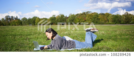 Leisure and people. Happy young woman lying in park on blanket, using laptop, relaxing outdoors and watching videos online on computer 108289426