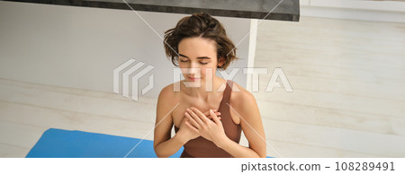 Sukhasana Hands To Heart. Portrait of young brunette fitness woman, girl holds hans on chest, doing yoga at home in sport leggins and bra, sitting on floor indoors 108289491