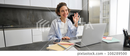 Happy young woman, tutor teacher students online. Girl connects to remote work meeting from her home laptop, says hello, waves hand at computer 108289500