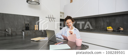 Portrait of young beautiful woman, studies at home, flips her workbook, works from home, uses laptop to learn on remote, makes notes during webinar 108289501