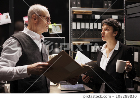 Senior private investigator showing criminology report files to investigator, discussing crime scene evident in arhive room. Police officer working at criminal case, analyzing suspect detalies Senior private investigator showing criminology report files to investigator, discussing crime scene evident in arhive room. Police officer working at criminal case, analyzing suspect detalies 108289830