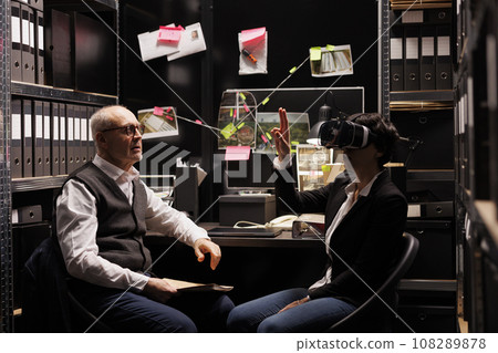 Private investigator wearing virtual reality goggles, using headset for crime scene simulation in arhive room. Police officers working overhours at criminal investigation case. Techology concept 108289878