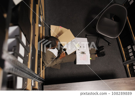 Criminologist drinking coffee and reading crime case on floor. Police investigator holding mug and folder with report papers while studying archival records at night time top view Criminologist drinking coffee and reading crime case on floor. Police investigator holding mug and folder with report papers while studying archival records at night time top view 108289889