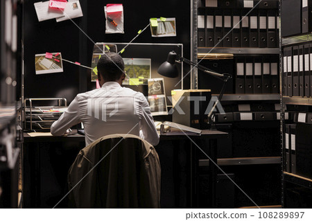 Detective sitting at desk and analyzing evidence scheme hanging on wall. Police law enforcement professional solving crime, studying clues and crime scene photos on board at night time Detective sitting at desk and analyzing evidence scheme hanging on wall. Police law enforcement professional solving crime, studying clues and crime scene photos on board at night time 108289897