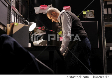 Private detective analyzing mysterious crime scene evidence, working overtime at solution to catch suspect. Elderly police officer reading confidential federal information in arhive room Private detective analyzing mysterious crime scene evidence, working overtime at solution to catch suspect. Elderly police officer reading confidential federal information in arhive room 108289916