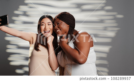 Two skincare models taking pictures on smartphone, using mobile phone to have fun with photos. Young beautiful women promoting self love and body positivity, self acceptance in studio. Two skincare models taking pictures on smartphone, using mobile phone to have fun with photos. Young beautiful women promoting self love and body positivity, self acceptance in studio. 108289937