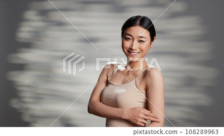 Beauty model using nourishing cream on shoulders, applying moisturizer on body to promote self acceptance and skincare routine. Luminous young woman advertising self love and cosmetics. 108289963