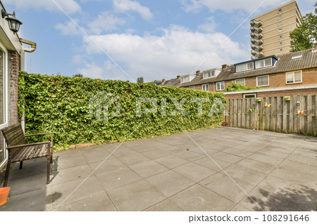 a backyard area with a fence, bench and some plants on the side of the house there is a blue sky in the background 108291646