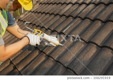 Worker man using silicone sealant adhesive  to fix crack of the old tile roof. 108291919