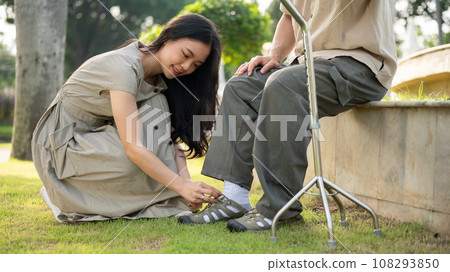 A caring Asian daughter is helping her senior dad put shoes on while relaxing in a park together. A caring Asian daughter is helping her senior dad put shoes on while relaxing in a park together. 108293850