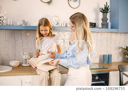 Blonde woman with little girl reading a book while standing at the kitchen. Family preparation holiday food. Mother and daughter is going to bake cookies. 108296735