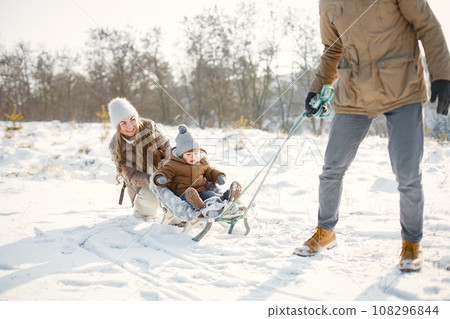 Portrait of happy family having fun on winter walk. Father carry his son on a sled. Family wearing warm jacket and knitted hats. 108296844