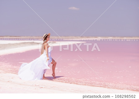 Woman in pink salt lake. She in a white dress and hat enjoys the scenic view of a pink salt lake as she walks along the white, salty shore, creating a lasting memory. 108298502