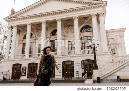 Woman street lifestyle. Image of stylish woman walking through European city on sunny day. Pretty woman with dark flowing hair, dressed in a beige raincoat and black, walks along the building. 108298505