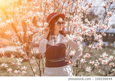 Magnolia park woman. Stylish woman in a hat stands near the magnolia bush in the park. Dressed in white corset pants and posing for the camera. 108298644