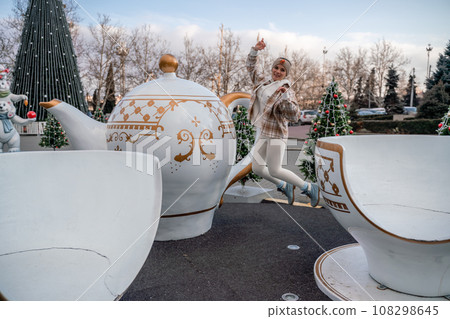 Woman Christmas Square. She stands near a large white cup, dressed in a light suit. With trees decorated with Christmas tinsel in the background Woman Christmas Square. She stands near a large white cup, dressed in a light suit. With trees decorated with Christmas tinsel in the background 108298645