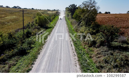 Looking down a gravel road with dust being blown by a vehicle. Path lined by power lines and trees 108299387