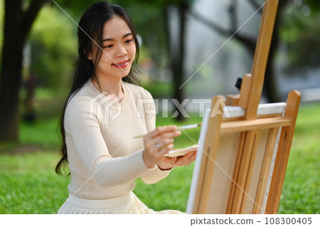 Charming young woman artist holding painting a picture on an easel at the park 108300405