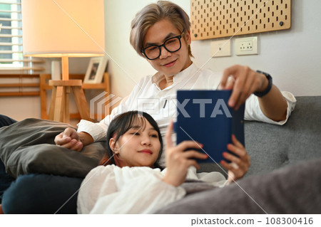 Loving young couple reading book while sitting together on sofa in living room 108300416