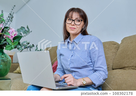 Mature woman sitting on living room sofa at home with laptop 108302178