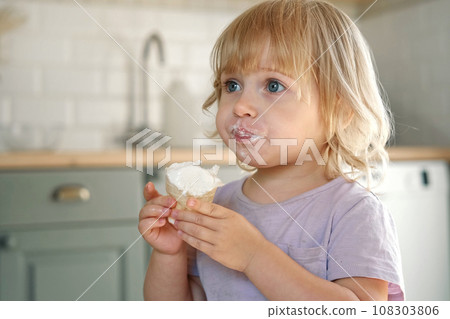 Baby girl enjoying ice cream. Pretty little toddler eating an ice-cream indoors, at home. Dining room background. Small child eats plombir and cream messy on her mouth. Cute kid with tasty sweet food. 108303806