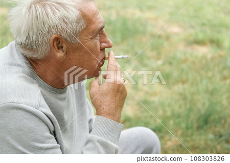 Senior man lighting up a cigarette outdoors against green nature background. Old thoughtful man smoking, looking aside. Close Up Face. Relaxed pensioner enjoy weekend rest outside. Unhealthy habits Senior man lighting up a cigarette outdoors against green nature background. Old thoughtful man smoking, looking aside. Close Up Face. Relaxed pensioner enjoy weekend rest outside. Unhealthy habits 108303826