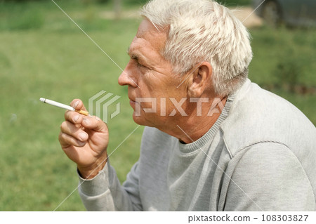 Senior man lighting up a cigarette outdoors against green nature background. Old thoughtful man smoking, looking aside. Close Up Face. Relaxed pensioner enjoy weekend rest outside. Unhealthy habits Senior man lighting up a cigarette outdoors against green nature background. Old thoughtful man smoking, looking aside. Close Up Face. Relaxed pensioner enjoy weekend rest outside. Unhealthy habits 108303827