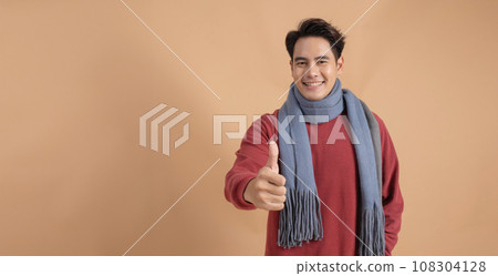 Happy and positive Asian man in red sweater and scarf giving thumbs up with ad empty space isolated on beige color background. Happy and positive Asian man in red sweater and scarf giving thumbs up with ad empty space isolated on beige color background. 108304128
