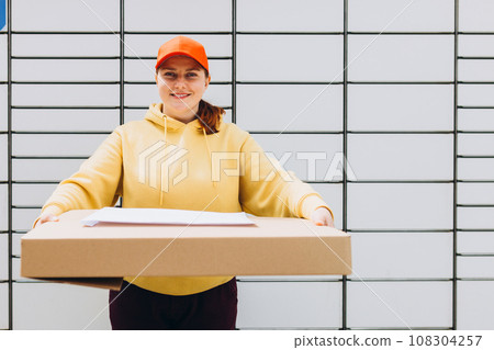 Happy 30s delivery woman in red hat giving packages. Concept of fast delivery to automatic self lockers 108304257