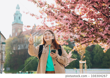Attractive young female tourist is exploring new city. Redhead 30s girl with smartphone resting near Wawel castle, Cracow, Poland. Traveling Europe in spring. High quality photo 108304267