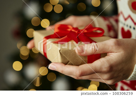 Woman in warm sweater holding gift box wrapping in craft paper with festive ribbon. Present for winter holidays. Christmas and Happy New Year celebrating 108306259