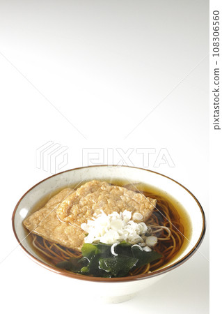 Hot fox soba noodles topped with two pieces of fried tofu, seaweed, and green onions are photographed against a white background. 108306560