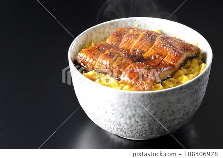 在黑色背景下拍攝的一碗熱氣騰騰的金子煎蛋捲和蒲燒鰻魚飯碗 108306978