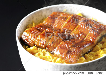 在黑色背景下拍攝的一碗熱氣騰騰的金子煎蛋捲和蒲燒鰻魚飯碗 108306985