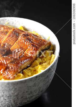 A piping hot rice bowl of kinshi omelet and kabayaki eel photographed against a black background 108306998