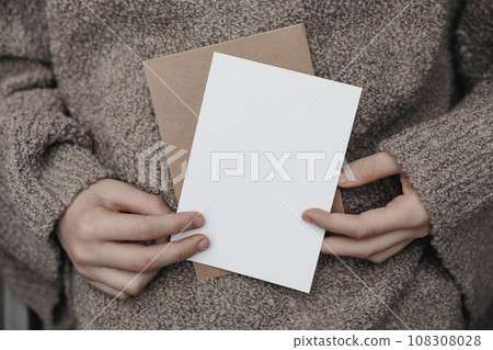 Winter, autumn cozy stationery still life. Child, female hands in beige pullower holding blank greeting card, craft envelope. Invitation mockup on knitted wool sweater. Blurred background. Winter, autumn cozy stationery still life. Child, female hands in beige pullower holding blank greeting card, craft envelope. Invitation mockup on knitted wool sweater. Blurred background. 108308028