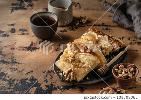 Crepes or thin pancakes with honey and nuts on brown plate over wooden background. Delicious homemade crepes for breakfast dark moody, text space 108308065