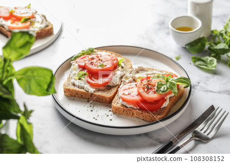 Sandwiches or toasts with tomatoes, cream cheese, olive oil and basil on a plate on white marble background. Traditional italian mediterranean food 108308152