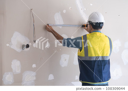 Skilled worker standing near shabby wall measuring distance with tape-line before drilling holes man in protective hardhat and vast renovates living room interior 108309044